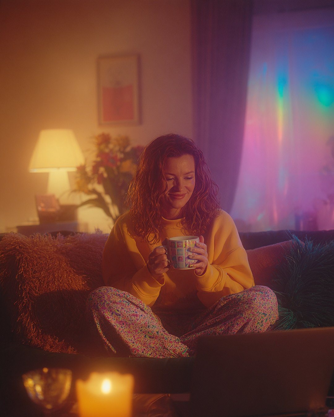 a woman at home, curled up on her sofa, holding a cup, in the light of a lamp