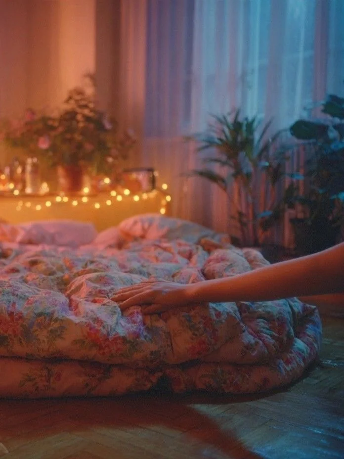 a hand smoothing a folded floral eiderdown on the floor, in a prepared bedroom, among fairy lights and plants