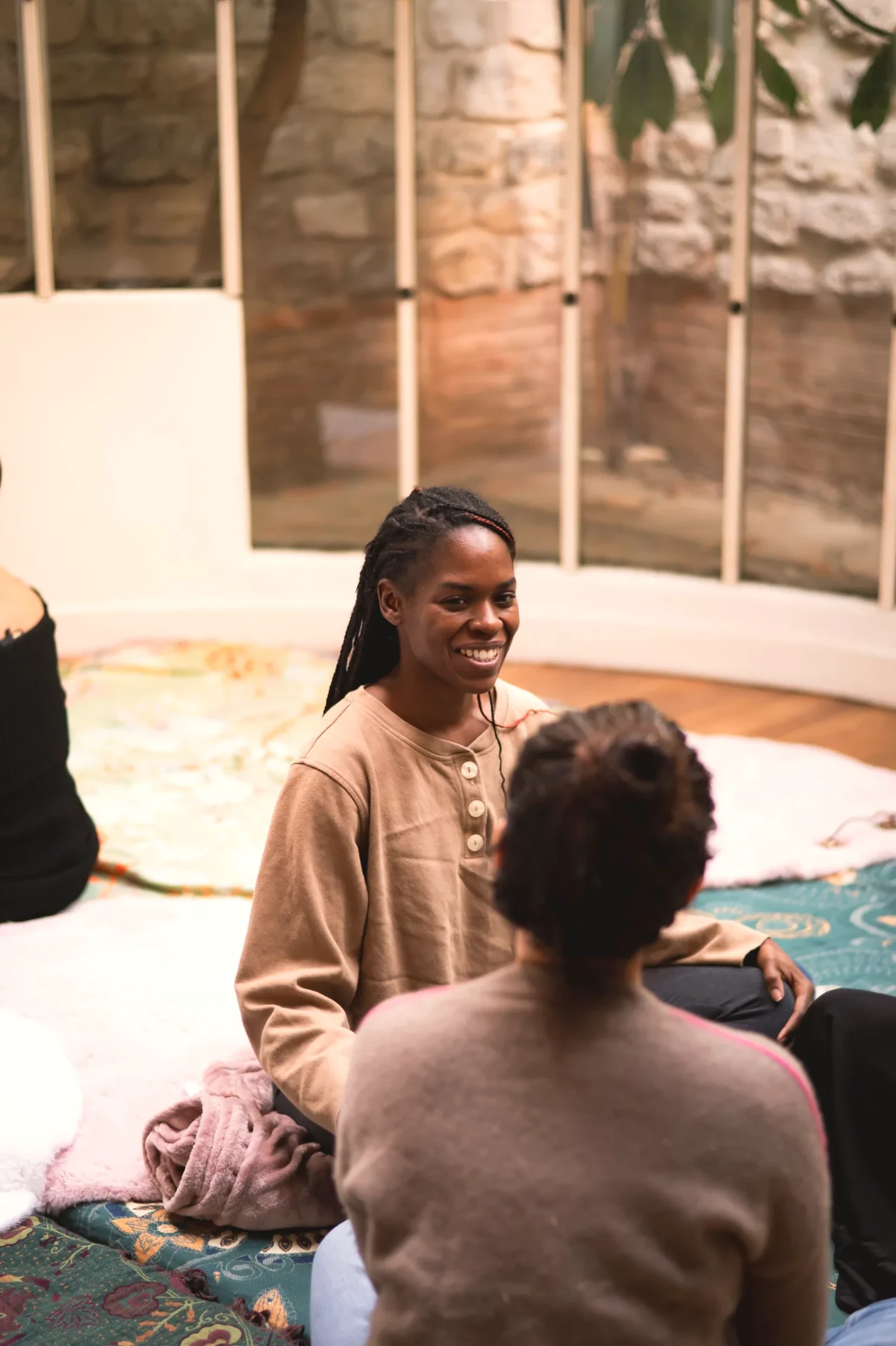 two women face to face on a rug, one listening with a smile, a stone wall behind