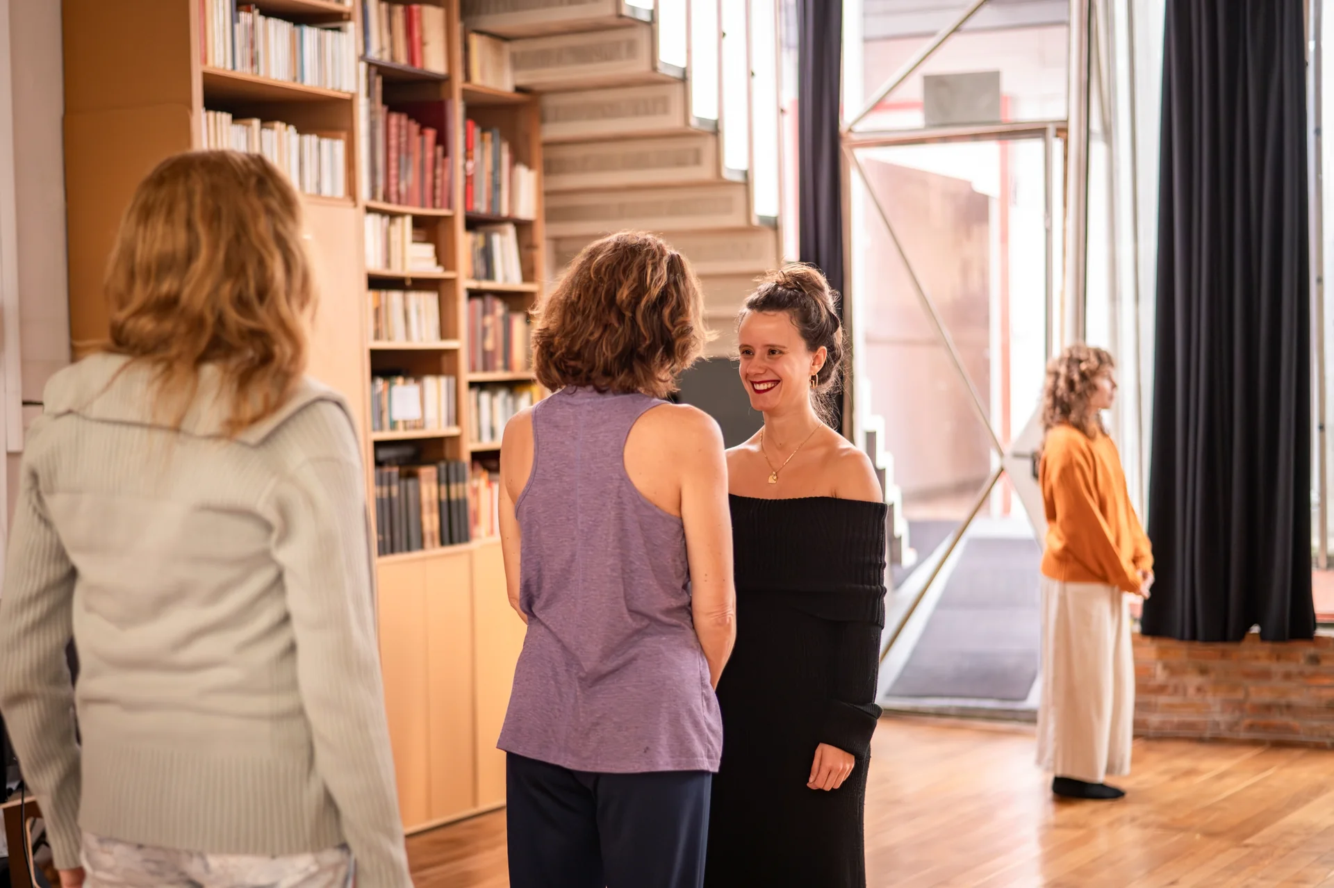 in a library, four women stand talking; one smiles at the woman speaking to her, another comes in through the glass door